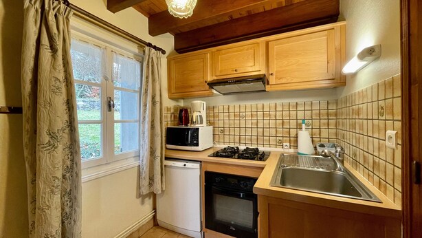 A compact kitchen with wooden cabinets, a microwave, a stove, a dishwasher, and a window with curtains. - Chalet familial près de la forêt avec grand terrain, jeux pour enfants et cheminée (Araules)