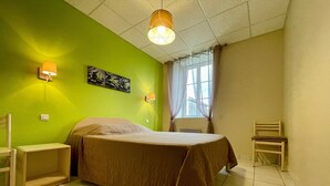 Une pièce comprenant un lit, deux chaises, une fenêtre avec des rideaux et une lampe fixée au mur.