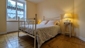 A bedroom with a wooden floor, a bed with a floral bedspread, a bedside lamp, and a window with curtains. - Stunning 2BR Apartment in Historic Le Puy-en-Velay with Modern Amenities (Le Puy-en-Velay)
