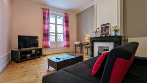 A living room with a black sofa, a flat-screen TV, a fireplace, and a dining area with a table and chairs. - Stunning 2BR Apartment in Historic Le Puy-en-Velay with Modern Amenities (Le Puy-en-Velay)