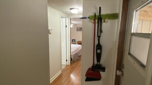 A narrow hallway with wooden flooring, a door on the right, and a bed visible in the background.
