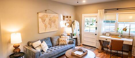 A living room with a sofa, coffee table, and a desk with a chair. There is a ceiling fan, a world map on the wall, and a window with curtains.