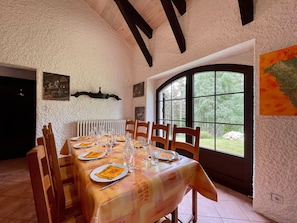 A dining area with a table set for six, wooden chairs, a white textured wall, a black ceiling beam, and a window with a view of greenery. - Tranquil Getaway: Cozy Home with Forest Views, Fireplace, and Family Amenities (Queyrières)
