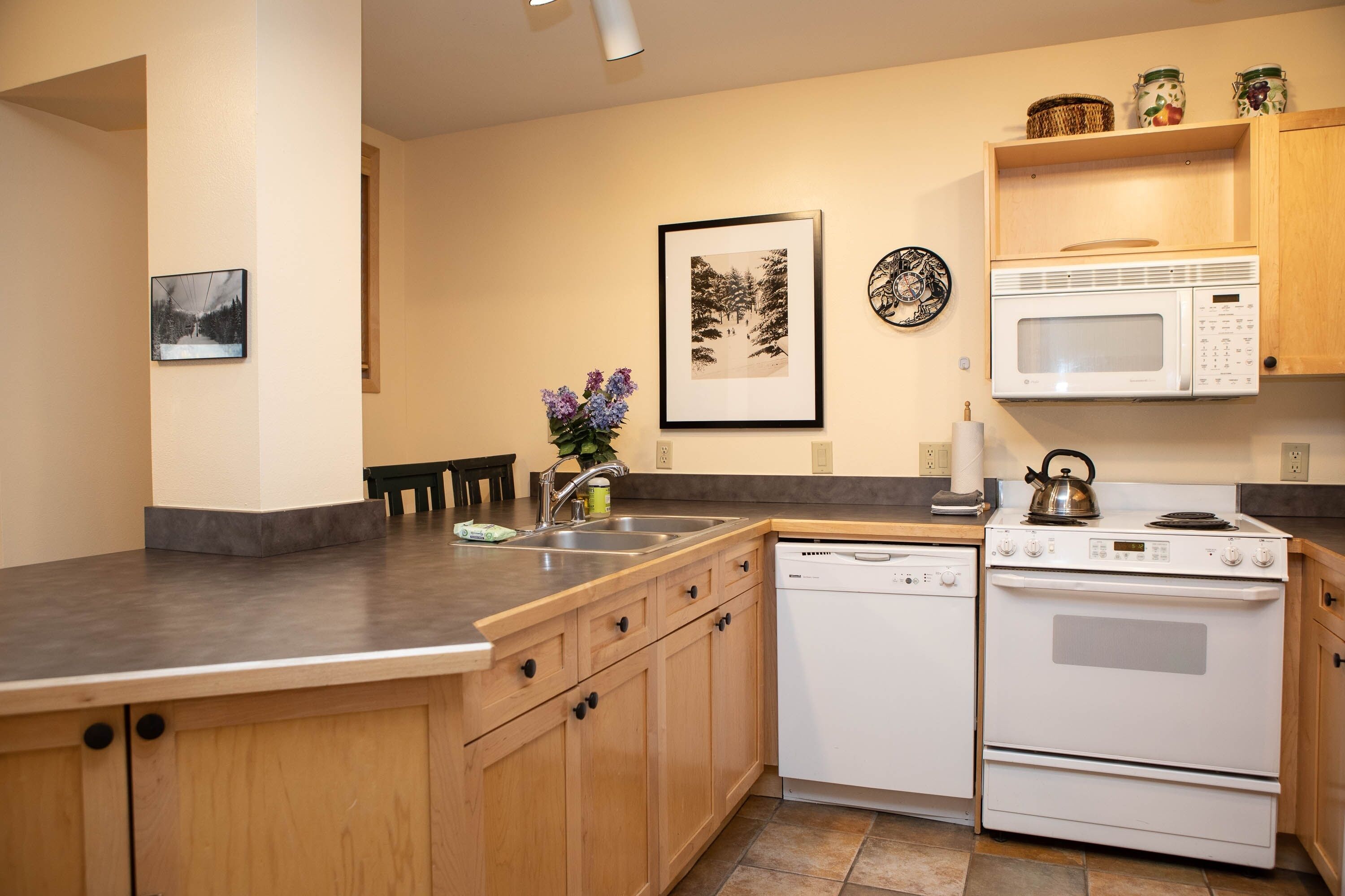A kitchen with wooden cabinets, a white dishwasher, a white stove, and a microwave. There are framed pictures on the wall and a vase with flowers on the counter.