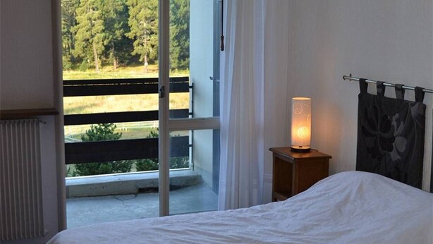 A bedroom with a bed, a nightstand with a lamp, a window with a view of trees, and a curtain. - Cozy 2-Room Apartment with Cabin for 6, Near Shops and 800m from Ski Slopes (Bolquère-Superbolquère)