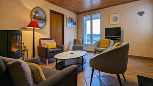 A cozy living room with a fireplace, comfortable seating, a television, and a large window offering a view of the outdoors. - Cottage with private terrace and garden in Saugues, all inclusive (Saugues)