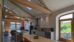 Una cocina moderna con vigas de madera, una zona de comedor con mesa de madera y sillas rojas, y vista a un paisaje verde a través de un amplio ventanal.