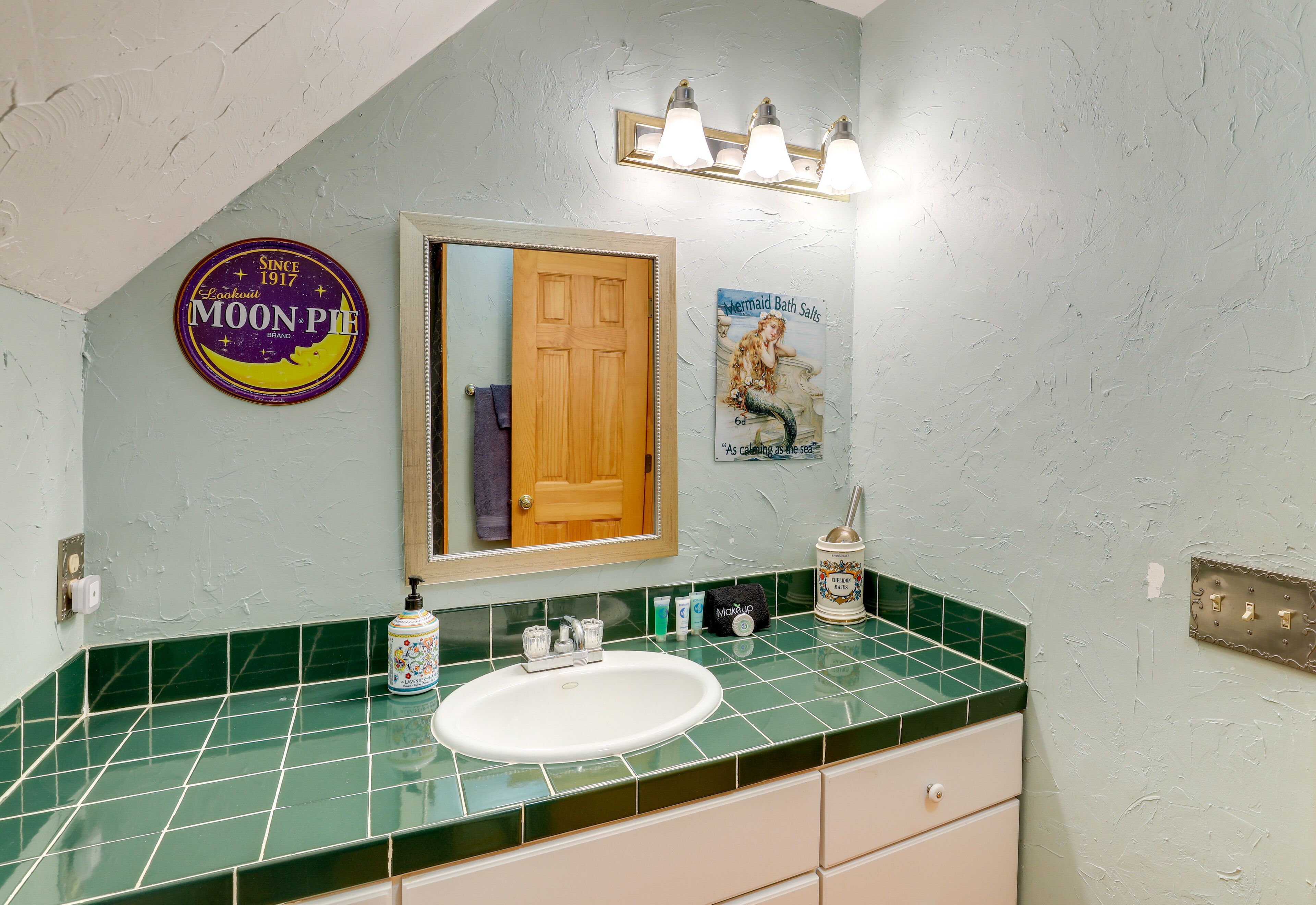 A bathroom with a green tiled countertop, a white sink, a mirror, a wooden door, and a light fixture above the mirror.