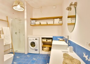A bathroom with a glass shower, a washing machine, a sink, and shelves with towels.