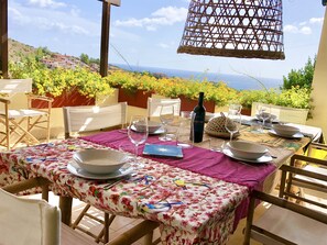 An outdoor dining area with a table set for four, surrounded by chairs, and overlooking a scenic view of the sea and hills.