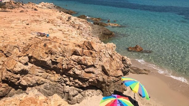 Ein felsiger Strand mit klarem türkisblauem Wasser, einem sandigen Ufer und einigen Sonnenschirmen.