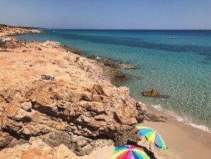 Uma praia rochosa com águas turquesa cristalinas, areal arenoso e alguns guarda-sóis.