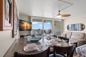 Uma sala de jantar com uma mesa posta para quatro pessoas, um ventilador de teto e uma pintura de um pôr do sol sobre a água.