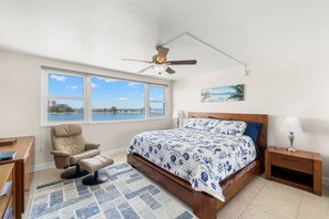 A bedroom with a bed, a desk, a chair, a nightstand, a lamp, a ceiling fan, a window with a view of the sea, and a painting on the wall.