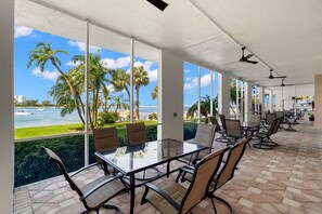 A spacious outdoor meeting area with a glass table and chairs, large windows offering a view of palm trees and the ocean.