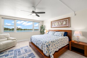 Ein Schlafzimmer mit einem Bett, einem Deckenventilator, einem Nachttisch, einer Lampe, einem gerahmten Bild und Blick auf das Meer durch große Fenster.