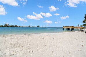 Ein Strand mit einer Holzsteganlage, die sich in ruhiges Wasser erstreckt, bewölkter Himmel und Gebäude in der Ferne.