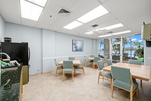 A modern dining area with light-colored walls, a large window with a view of palm trees and the sea, and several tables and chairs.