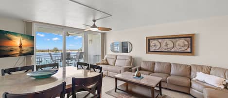 A spacious living area with a dining table, chairs, a sofa, and a coffee table. There is a ceiling fan, a painting of a sunset, and a framed picture on the wall.