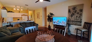 Uma sala de estar com uma mesa de jantar marrom escura, uma TV na parede e uma cozinha ao fundo.