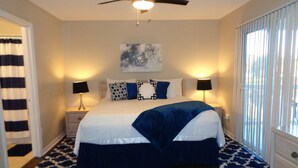 A bedroom with a bed, two bedside tables, a ceiling fan, a window with blinds, and a door with a striped curtain.