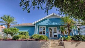 A two-story building with a blue exterior, a front entrance with a set of stairs, and a small garden area with palm trees and shrubs.