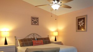 Un dormitorio con una cama, dos mesitas de noche con lámparas, un ventilador de techo, una obra de arte en la pared y un cuadro enmarcado.