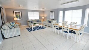 A spacious living room with a sofa, a dining table with chairs, and a coffee table. There are framed pictures on the walls and a window providing natural light. - Family-Friendly 1st Floor Poolside Condo | 3BR/2BA | 5 Miles to Disney! (Kissimmee)