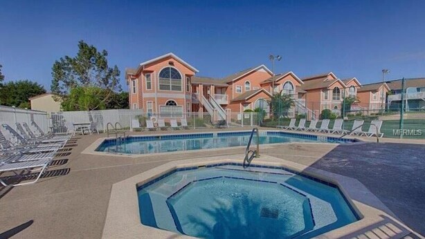 A swimming pool area with lounge chairs and a hot tub, surrounded by a building with a red and beige color scheme. - Family-Friendly 1st Floor Poolside Condo | 3BR/2BA | 5 Miles to Disney! (Kissimmee)