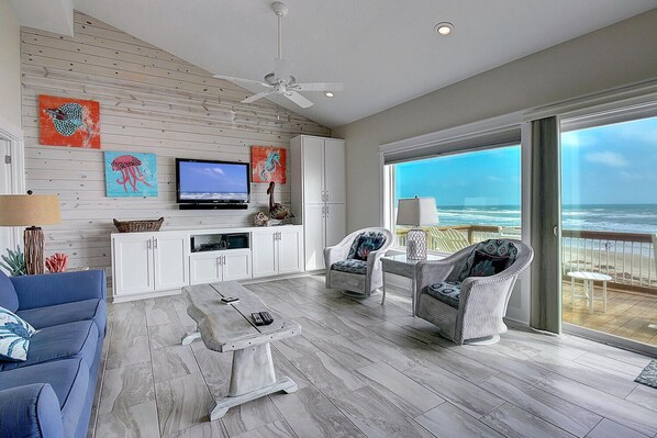 A spacious living room with a large TV, white cabinets, and a balcony with a view of the ocean.