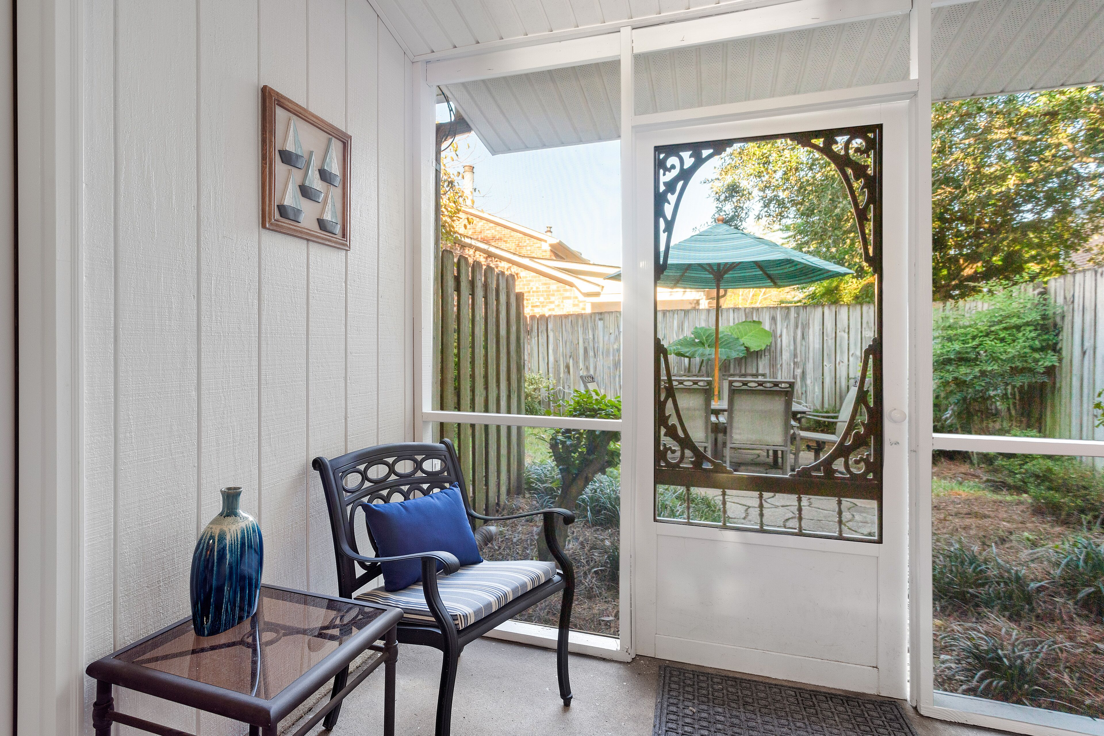 A covered porch with a chair and a table, a wooden fence, and a green umbrella in the background.