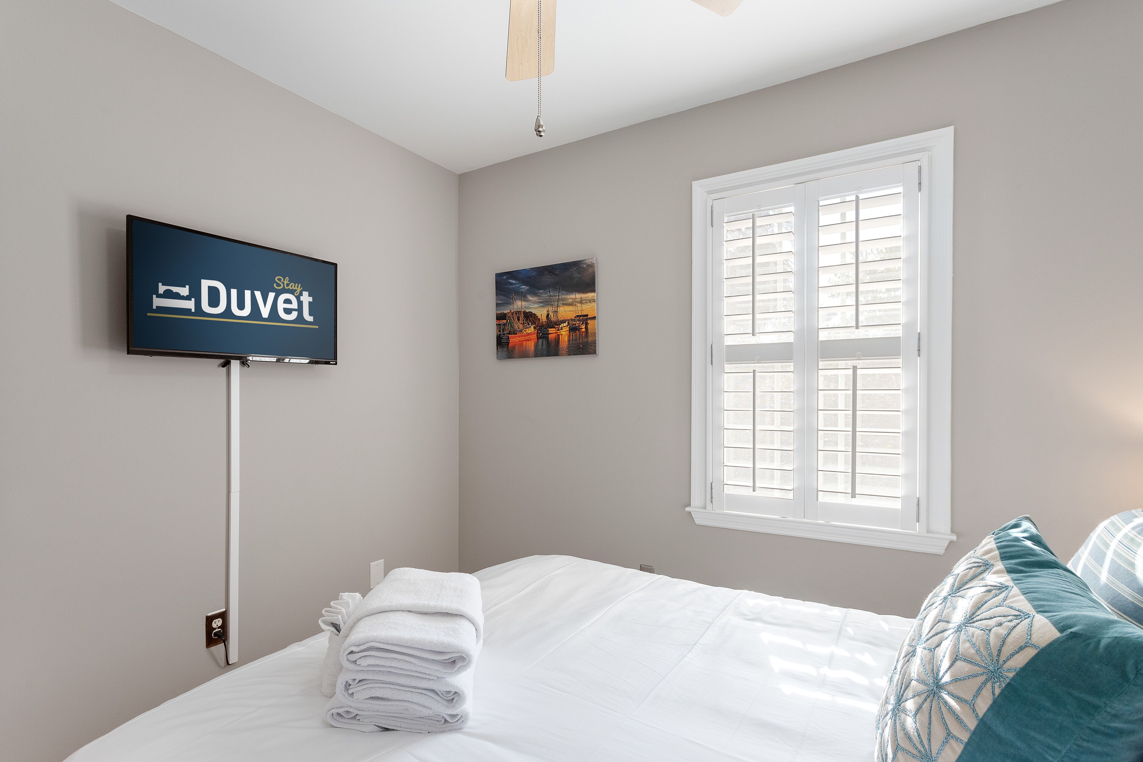 Habitación de hotel con cama, televisor montado en la pared, ventana con persianas y un cuadro en la pared.