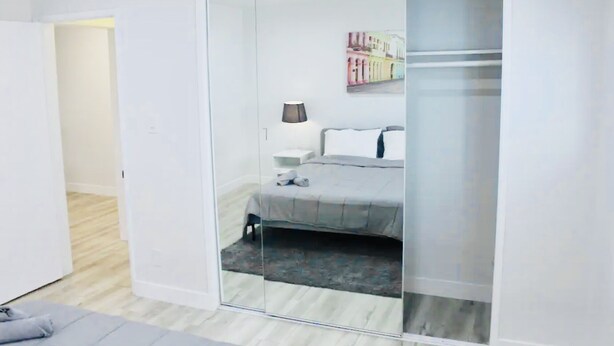A modern bedroom with a glass partition, a bed with grey bedding, a nightstand with a lamp, and a colorful artwork on the wall. - 1BR Apt in Culver City by Just Bring Your Toothbrush (Culver City)