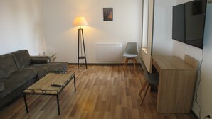 Uma sala de estar com sofá, mesa de centro, abajur, radiador, televisão e uma poltrona.