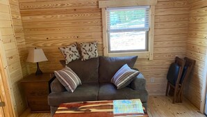 A cozy cabin interior with a wooden floor, a gray sofa with patterned cushions, a small table with a wooden top, and a window with blinds. -     Secluded Log Cabin • Trails • 7 Minutes to French Lick • Pet Friendly (French Lick)