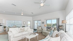 A spacious living room with a white sofa set, a coffee table, and a ceiling fan. There are large windows providing natural light and a view of the outdoors.