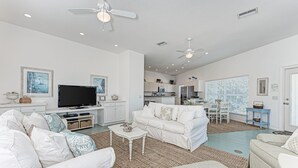 A spacious living room with a white sofa, a coffee table, and a TV. There is a kitchen area in the background with cabinets and a dining table.