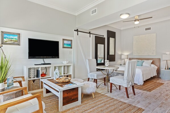 Une chambre d’hôtel moderne dotée d’une télévision à écran plat, d’un lit avec du linge de lit blanc, d’un coin repas avec une table et des chaises blanches, et d’une grande fenêtre.