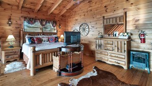 A wooden cabin interior with a bed, a round table, a dresser, and a fireplace.