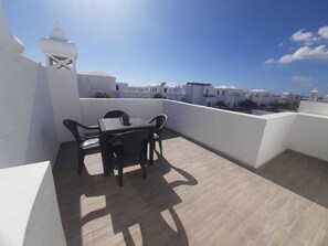 A rooftop terrace with a table and chairs, a potted plant, and a view of white buildings under a clear sky.