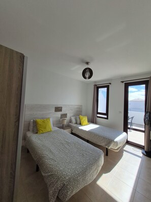 A room with two beds, a ceiling fan, a window with a view, and a wooden door.