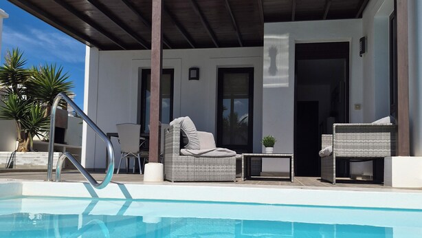 A modern pool area with a clear blue pool, a covered patio with wicker furniture, and a white building with large windows. - Villa with private heated swimming pool, Wifi, Prime video, BBQ, tennis table (Playa Blanca)
