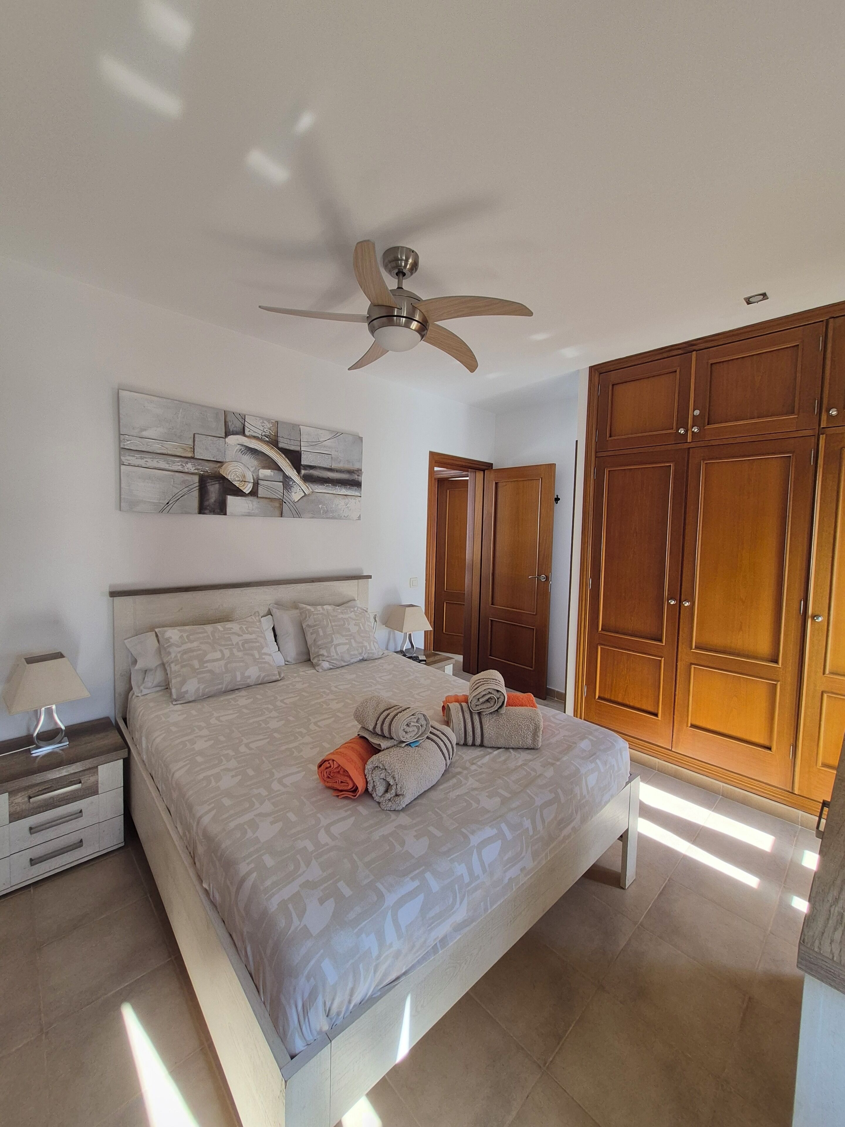 Een slaapkamer met een bed, een plafondventilator, een houten garderobe en een schilderij aan de muur.