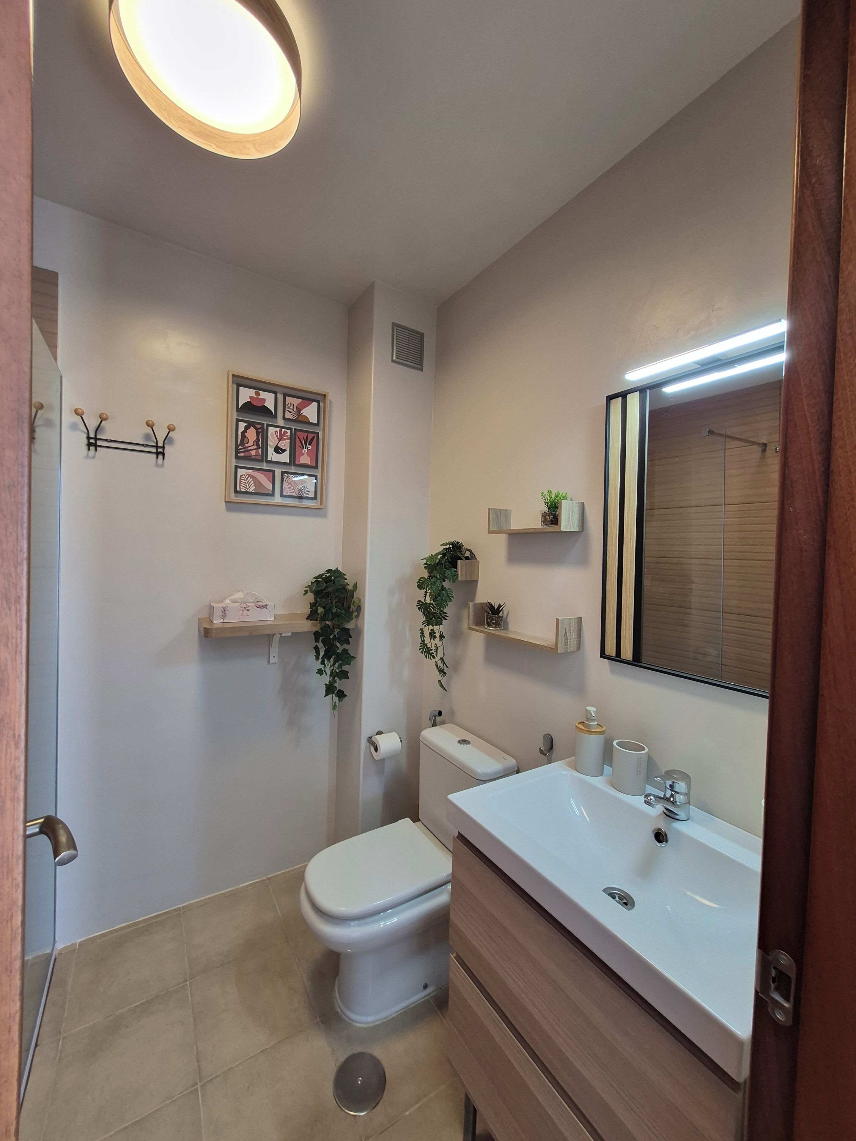 A modern bathroom with a toilet, sink, and mirror. There are wall-mounted shelves with plants and toiletries.