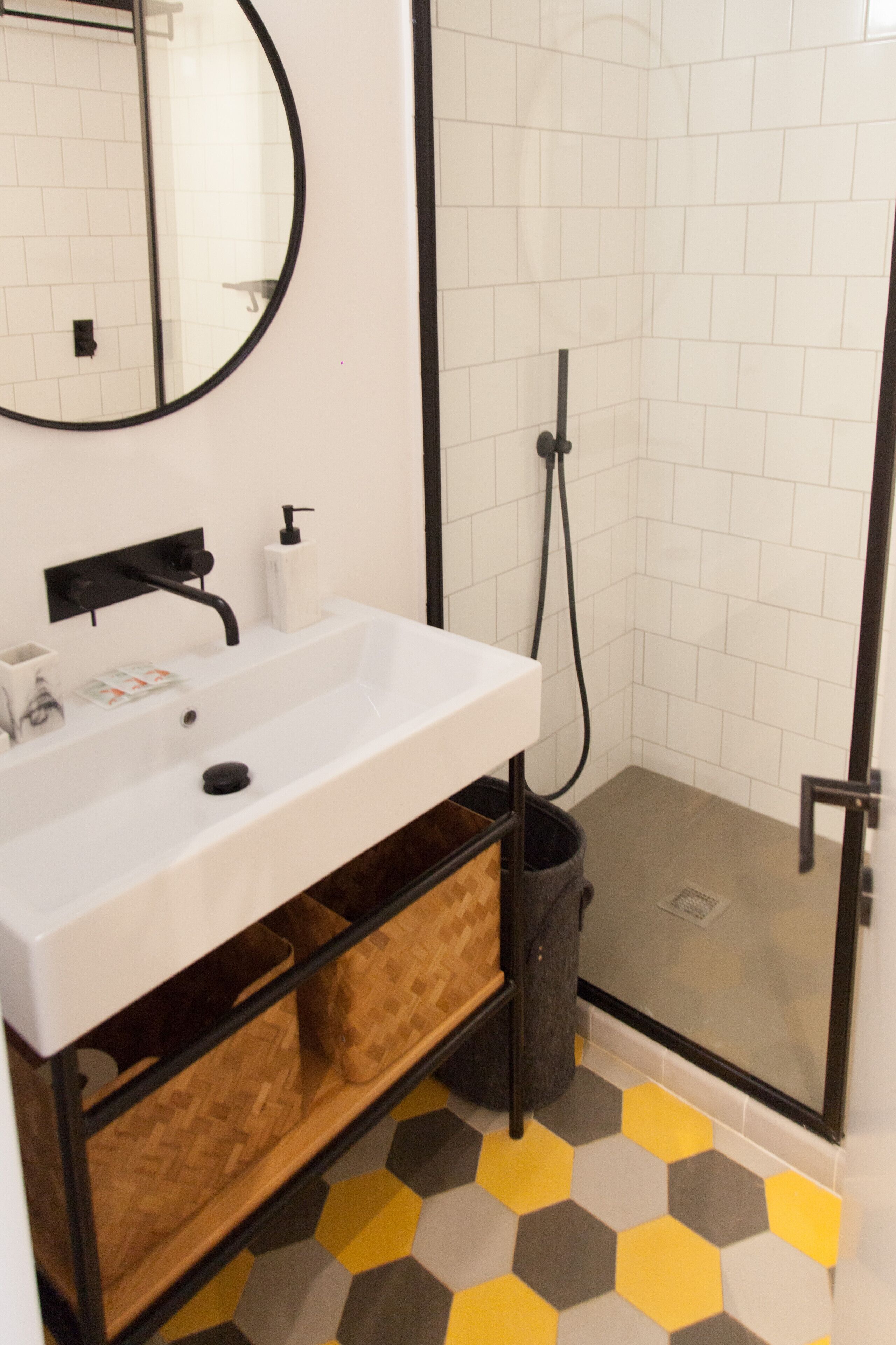 Een badkamer met een witte wastafel, een zwarte kraan en een spiegel. Er is een douchegedeelte met een glazen deur en een zwarte douchekop. De vloer heeft een geometrisch tegelpatroon.
