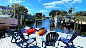 A wooden dock with kayaks and chairs, a canal with boats, and a clear sky. - Stunning Remodel, HEATED POOL, Brand New Furnishings, Full Canal Sunset View (Cape Coral)