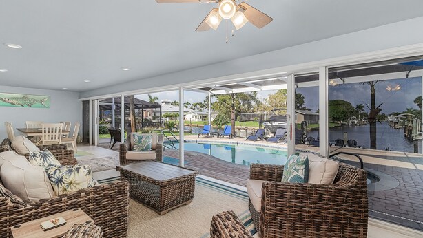 A spacious indoor area with wicker furniture, a ceiling fan, and a view of a swimming pool and outdoor seating. - Stunning Remodel, HEATED POOL, Brand New Furnishings, Full Canal Sunset View (Cape Coral)