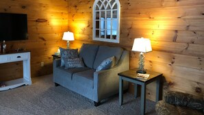 Ein gemütliches Zimmer mit Holzwänden, einem grauen Sofa, zwei Lampen, einem Fernseher und einem dekorativen Fenster.