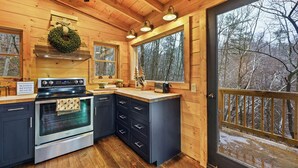 A wooden cabin kitchen with a stove, oven, and a large window overlooking a snowy forest. - Free Tickets | Wood Burning Fireplace, Hot Tub, WIFI, Pool Table, Sleeps 7 (Gatlinburg)