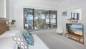 A bedroom with a large window, a bed with patterned pillows, a bedside table with a lamp, a TV on a wooden stand, and a framed picture on the wall. - Seascape Kiama: Ocean Views, Family Fun, Pet-Friendly Perfection managed by HAUS (Kiama)